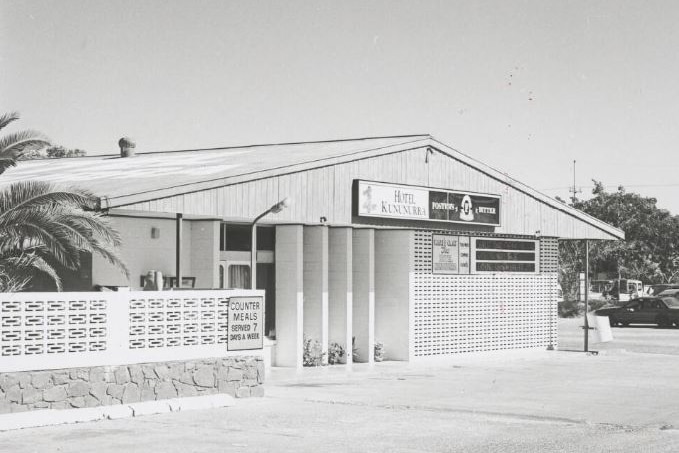 A black and white photo of a building with tin sloping roof, palm tree and sign on front.