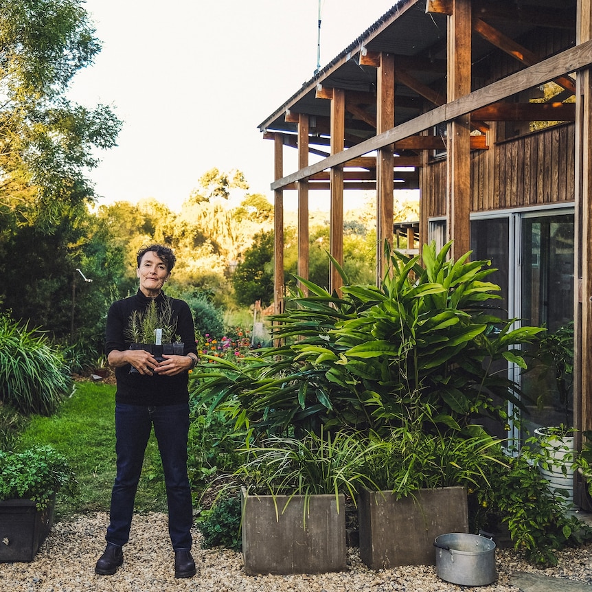 Edwina stands holding some small plants in her lush green garden.