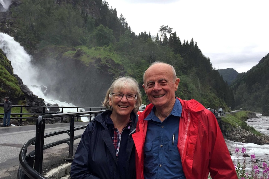 Julie and Jeff Wicks stand smiling in front of a waterfall. Behind them are forest covered mountains.