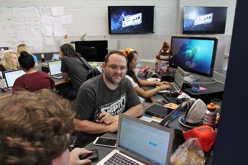 A young man sits at a desk surrounded by toys and other staff on computers