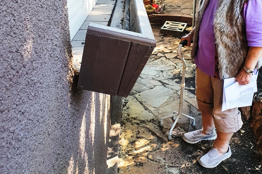 A woman with a walking stick stands in front of a brick window sill peeling off the wall.