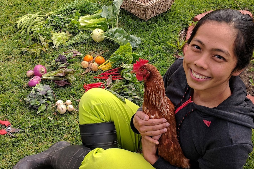 A woman holding a chicken sitting in the garden with her veggies produce smiling to the camera.