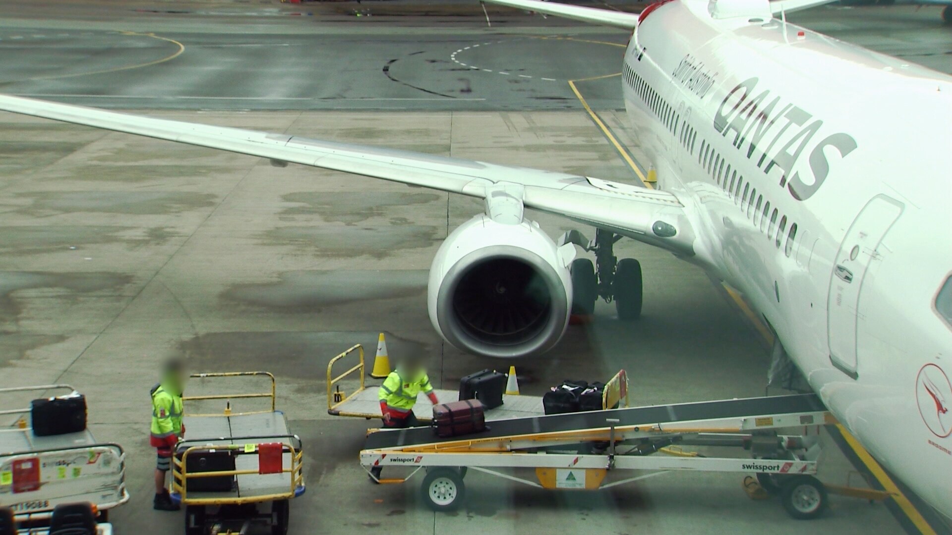 Baggage handlers loading luggage onto a conveyor belt beside a Qantas plane.