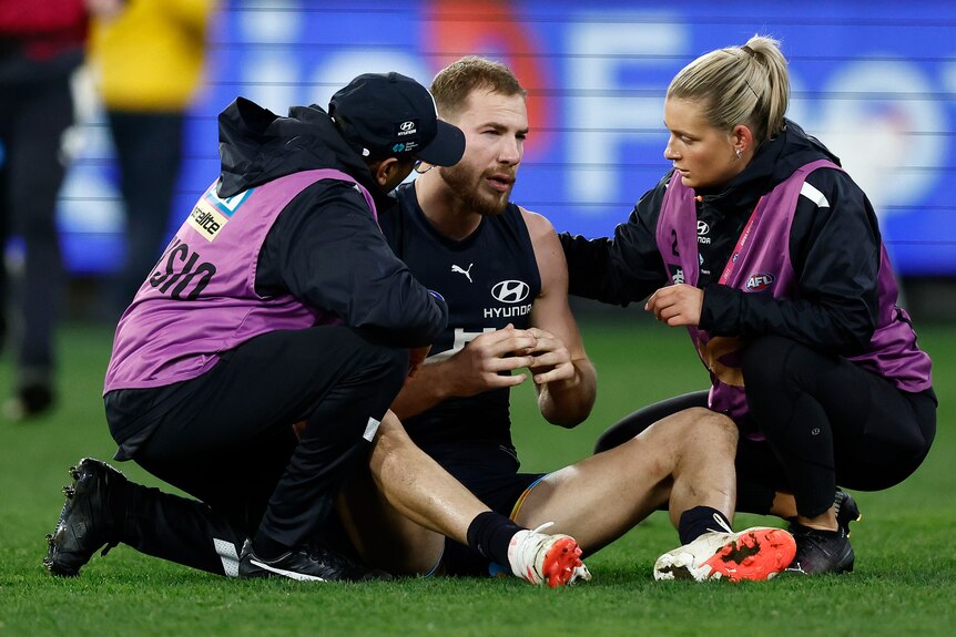 A Carlton AFL player sits dazed on the ground as two medical staff give him attention during a finals game.