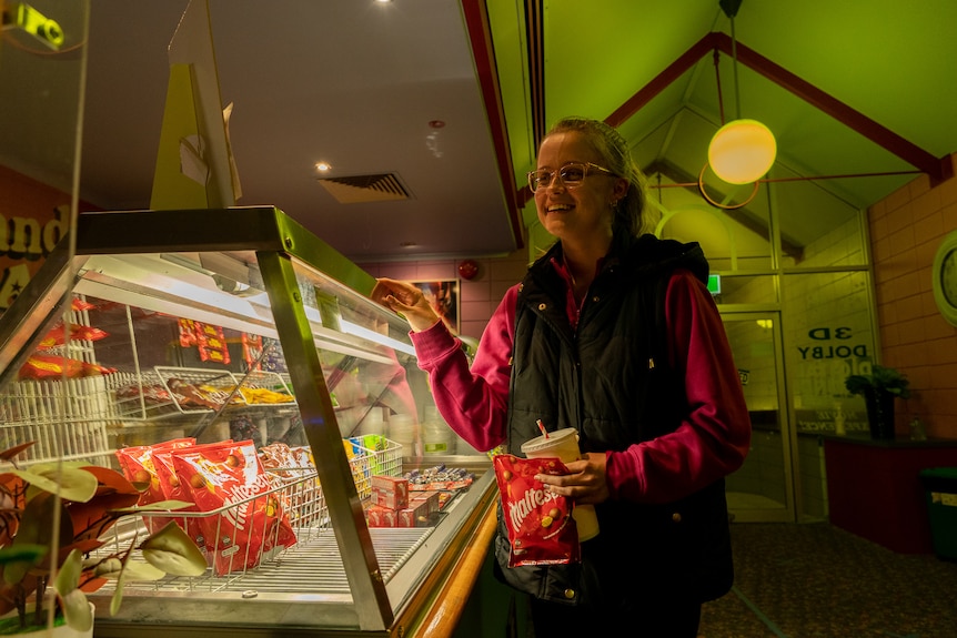 A young girl holding movie snacks chats across the candy bar to someone out of shot.