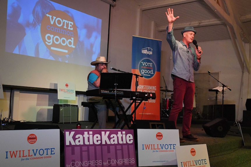 A man in a hat standing on stage with a microphone in front of a powerpoint presentation that says 'Vote Common Good'.