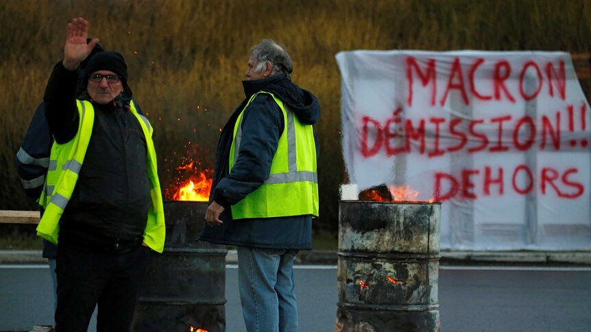 One protester in a yellow vest raises his hand with a sign in the background reading 'Macron Resign' in French.
