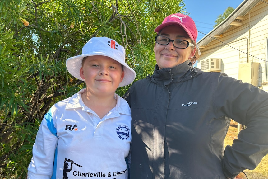 A boy in cricket gear and mum in jacket, red cap and reading glasses.