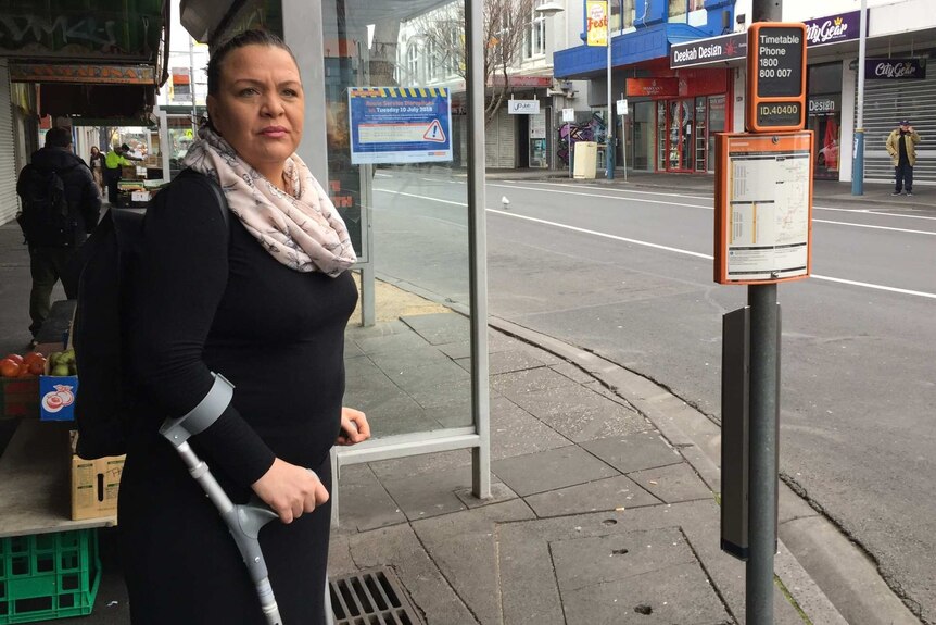 Dee Stanton, using crutches, waits at a bus stop in Footscray.