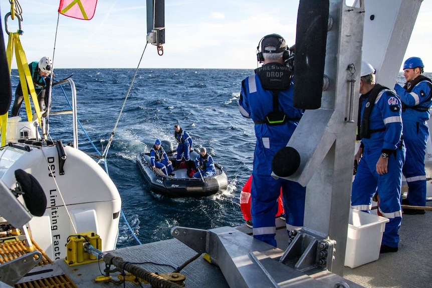scientists in blue uniforms stand on a boat and life boat in the ocean as a diver stands on a submersible