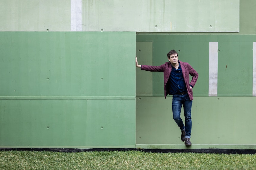 A white middle aged man wearing a maroon blazer, a blue shirt and jeans stands against a green wall.
