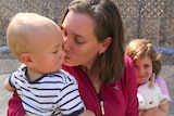 A woman in her backyard, kisses her young son while her daughter hugs a toy in the background.