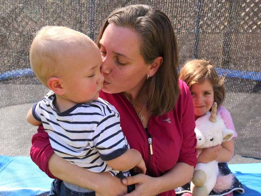 A woman in her backyard, kisses her young son while her daughter hugs a toy in the background.