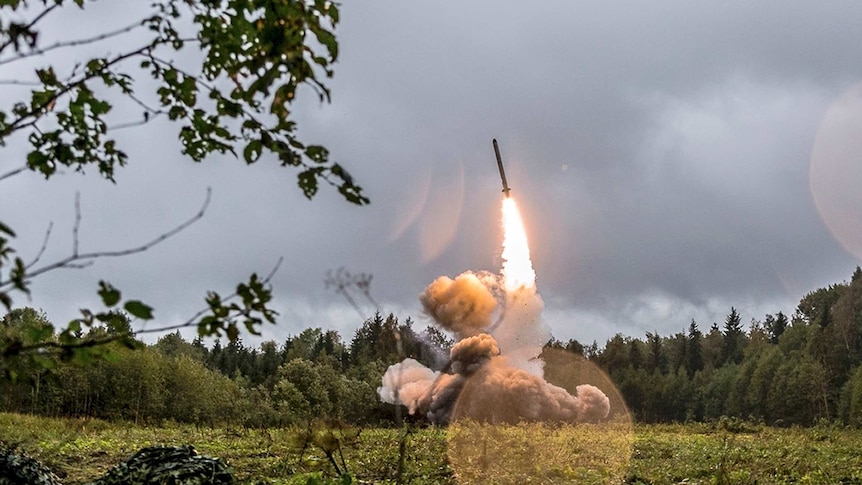 A Russian Iskander-K missile launches in a green field surrounded by trees.