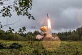 A Russian Iskander-K missile launches in a green field surrounded by trees.