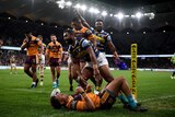 An NRL player stands up, gesturing after scoring a try, surrounded by opposition players.