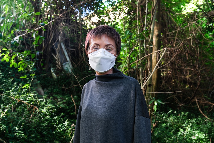 A woman wears a white face mask as she stands in a block of land with thick trees