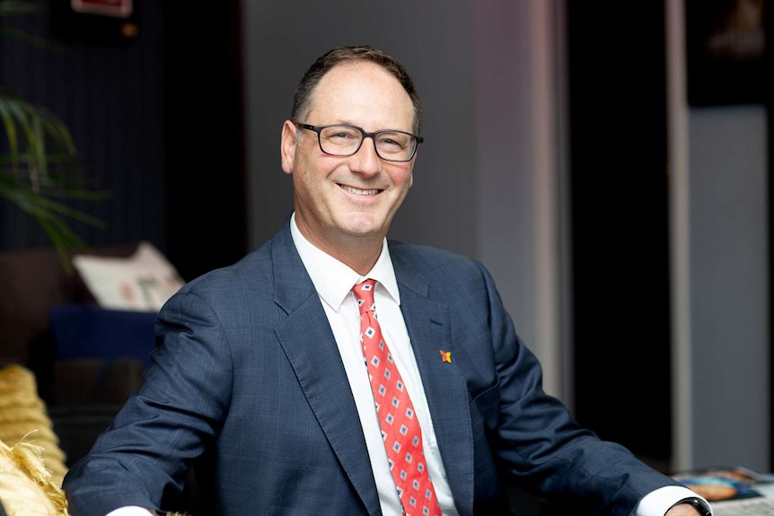 A man in a suit sits at a desk smiling at the camera.