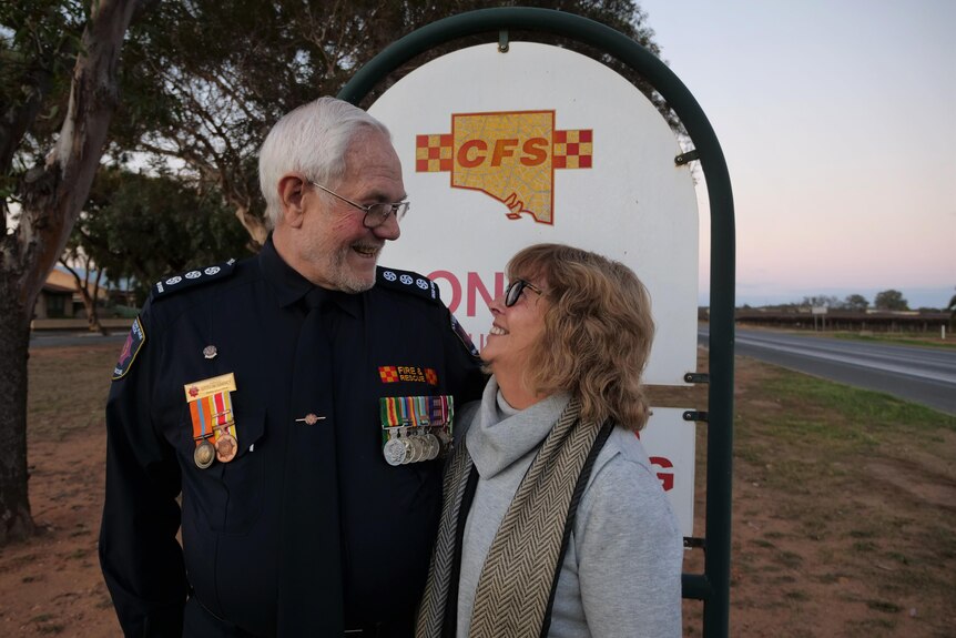 A senior man and woman look at each other with adoration. They stand in front of a fire service sign. The sun is setting.