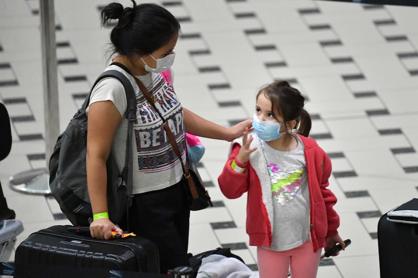 A woman puts her hand on the shoulder of a child wearing a red jumper. Both are wearing medical face masks wit suitcases nearby