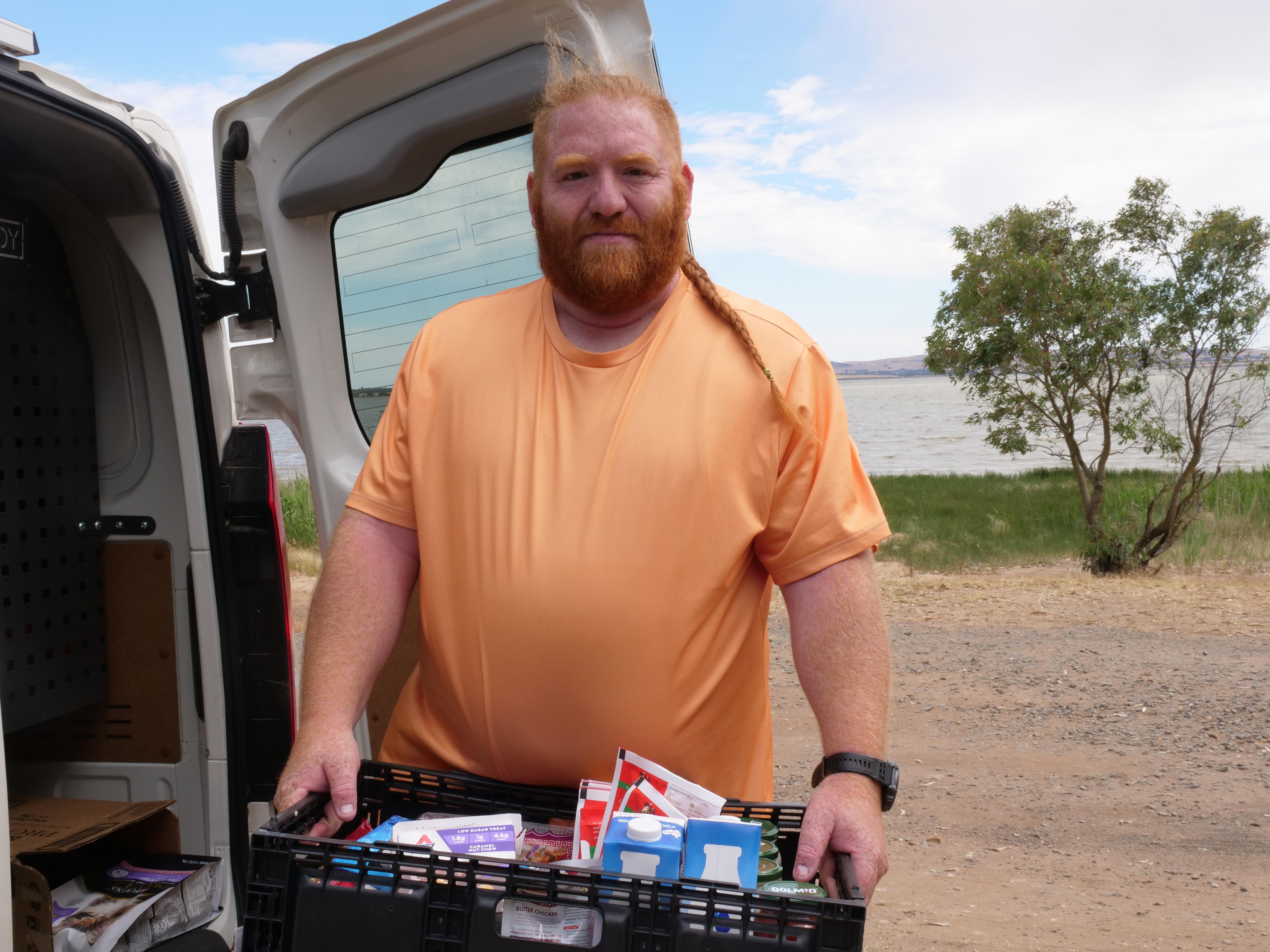 Hombre con camisa naranja sonriendo y sosteniendo una canasta de alimentos no perecederos frente a un lago. 