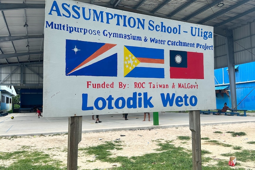 A sign in front of a covered basketball court that says 'multipurpose gymnasium and water catchment project'.