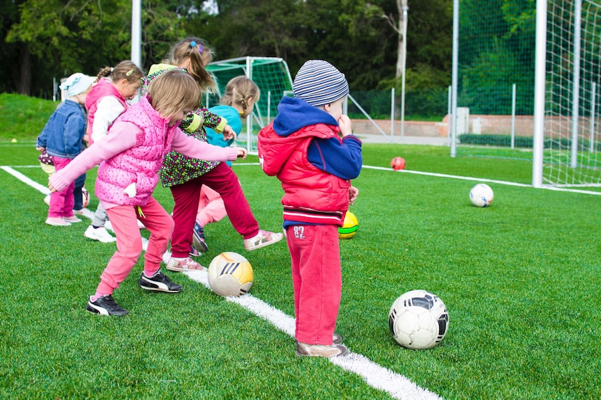 A line of small children on a football field kicking at balls to demonstrate how to teach kids to be good losers and winners.