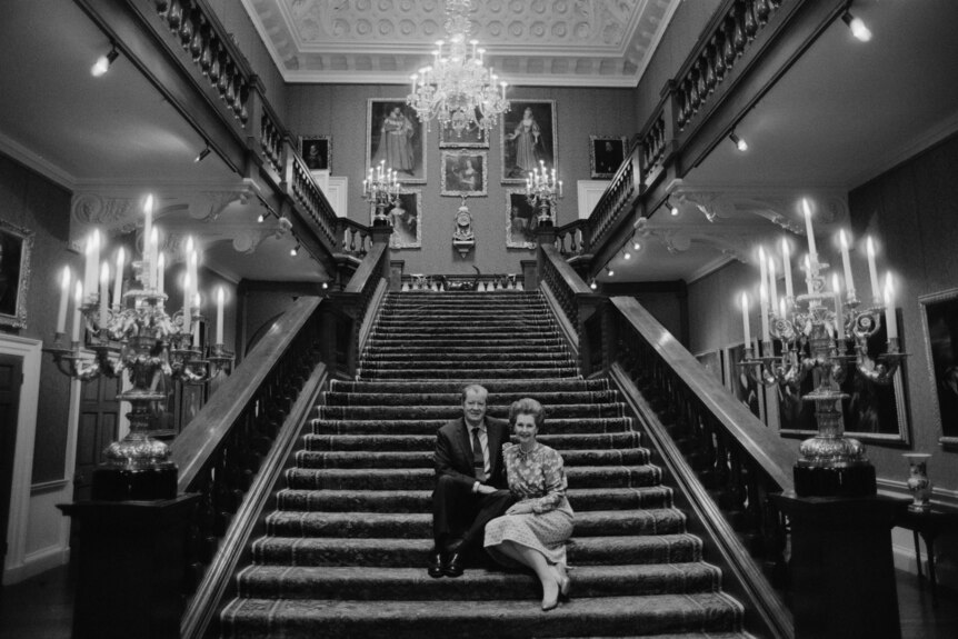 A man and a woman pose together at the bottom of a grand staircase