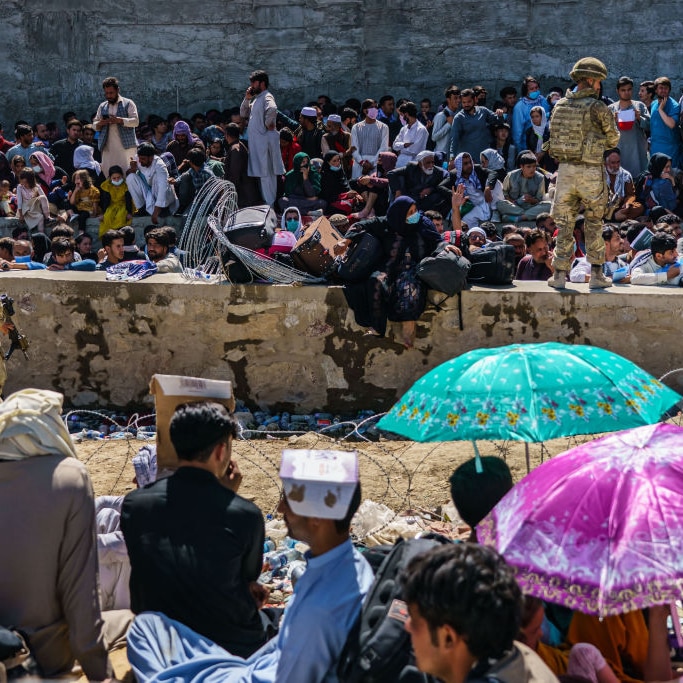 Crowd of people sitting in hot sun next to a canal and stone wall. Soldiers with guns