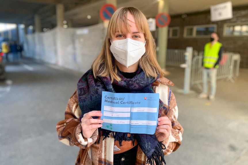 A young woman in a face mask holds up a medical certificate