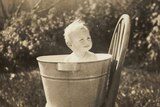 A black and white photo of a blonde baby sitting in a tin tub on a wooden chair.