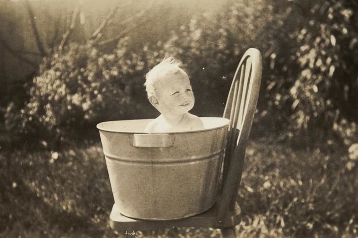A black and white photo of a blonde baby sitting in a tin tub on a wooden chair.