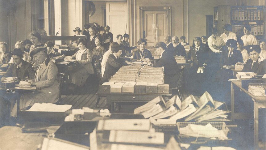An historic photo of women in suits, sitting at desks reading through large piles of cards and paperwork.
