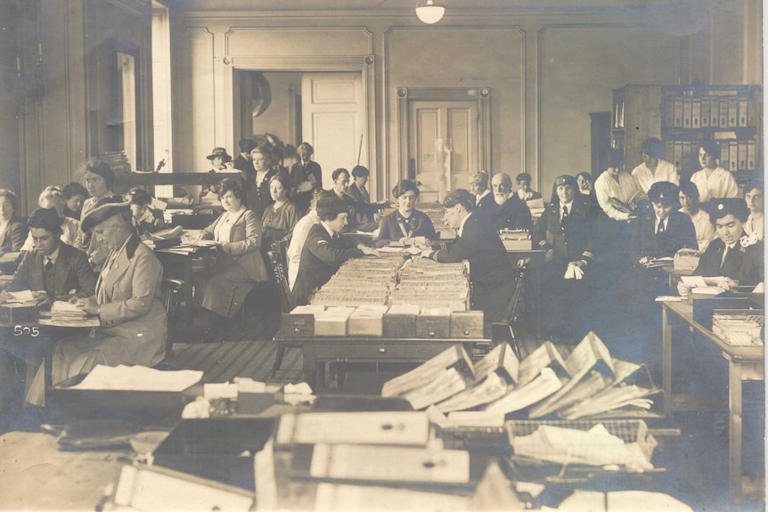 An historic photo of women in suits, sitting at desks reading through large piles of cards and paperwork.