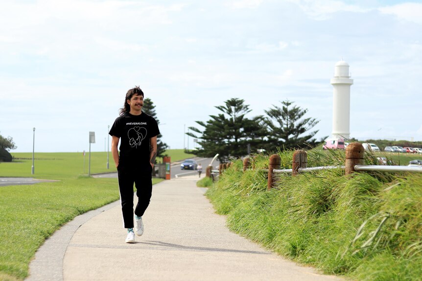 A man walks along a coastal footpath bordered by green grass wearing a black t-shirt and looking out to sea.