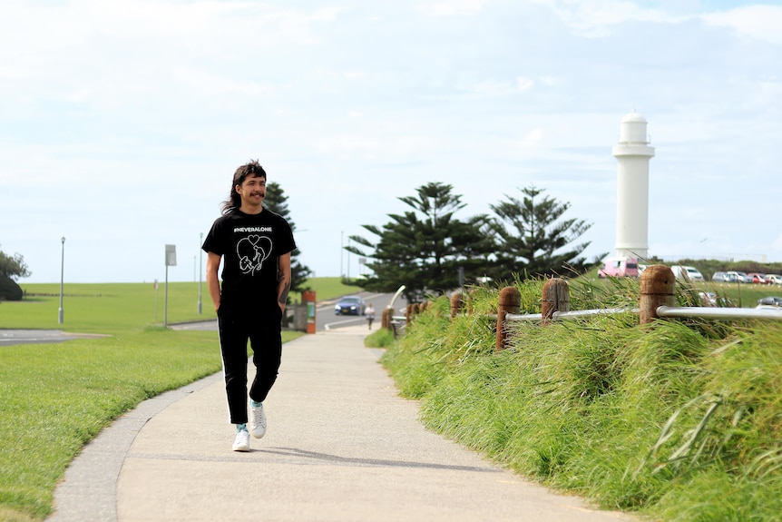 A man walks along a coastal footpath bordered by green grass wearing a black t-shirt and looking out to sea.
