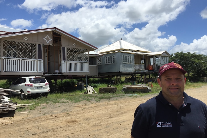 A man in a baseball cap stands in front of relocatable wooden homes.