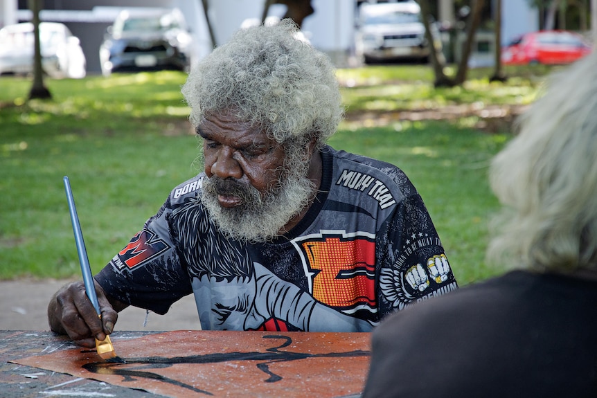 An Aboriginal man sits at an outside table painting on a piece of canvas. 