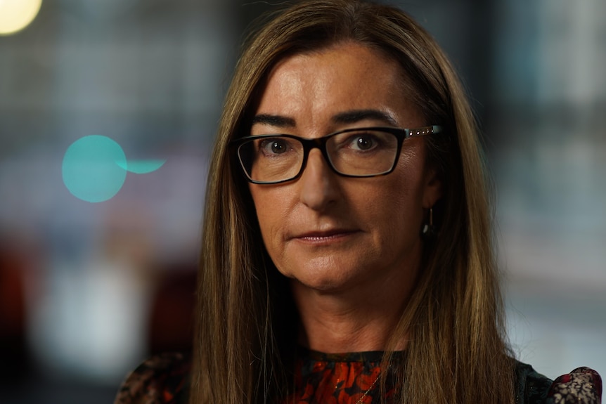 A portrait of a woman wearing glasses with dark blonde hair, serious facial expression