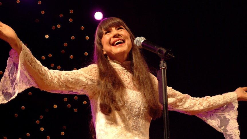 Judith wears a white long-sleeved dress and sings. She looks joyful.