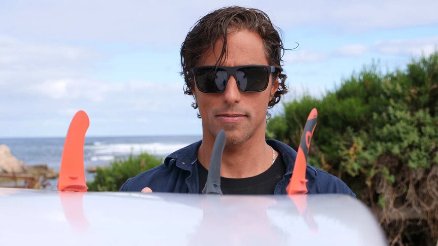 A man in sunglasses holds up a surfboard with the ocean in the background.