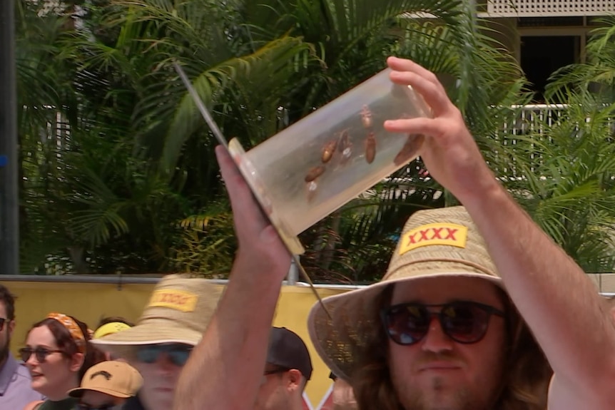 a man in a hat holds a clear cylinder full of cockroaches above his head