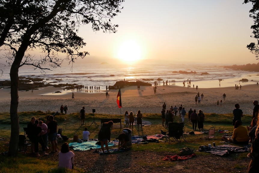 People gathered on a beach watching the sun rise with an Aboriginal flag