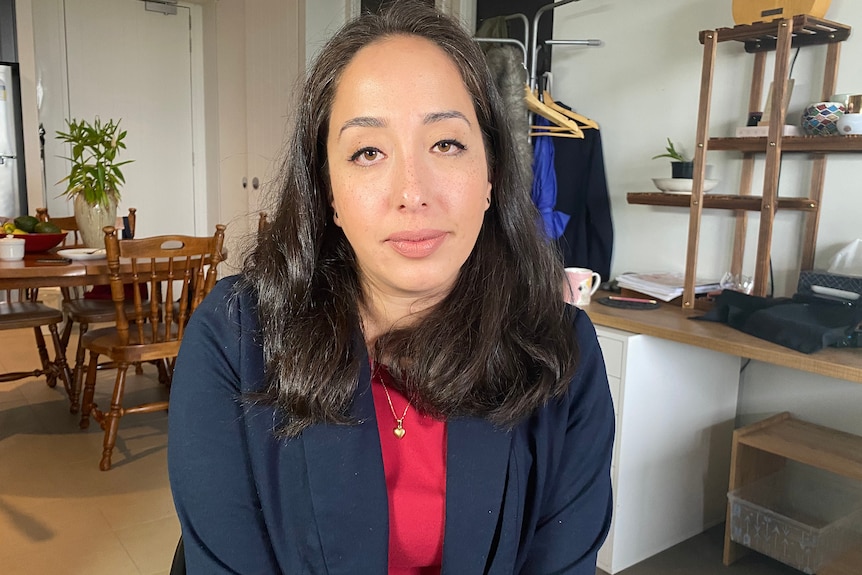 A woman with long dark hair wears a blue jacket over a red top.