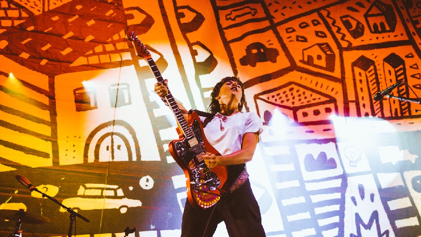 Courtney Barnett performing, playing guitar in front of large album artwork illustration