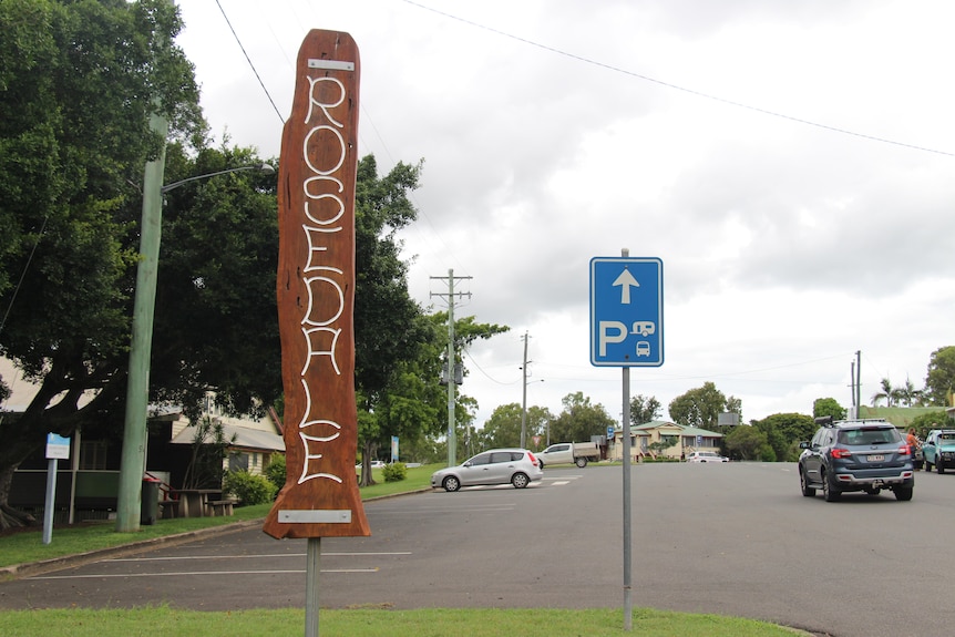Jalan utama kota pedesaan, beberapa mobil diparkir, papan kayu bertuliskan 'Rosedale'.