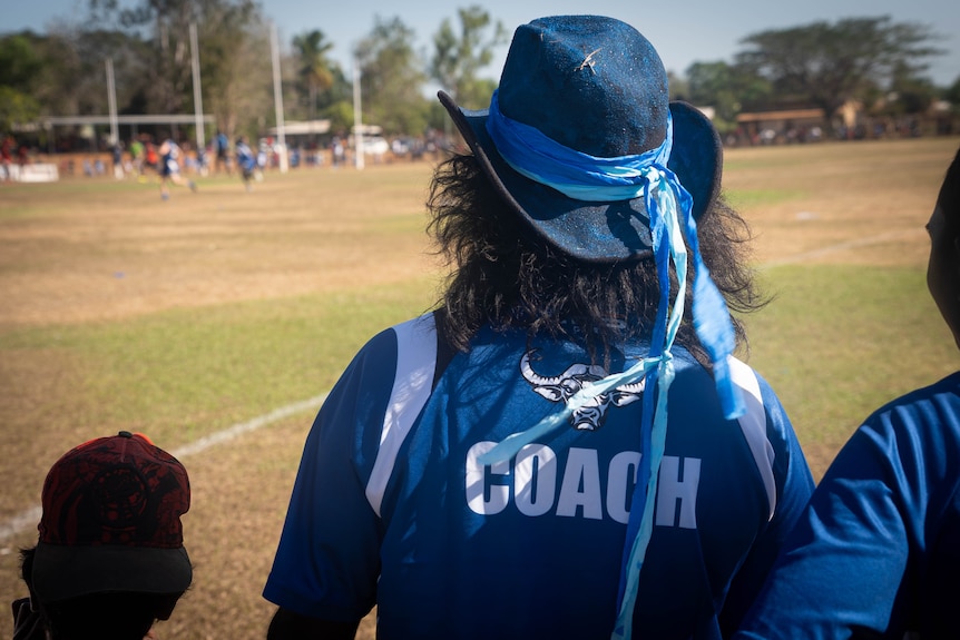 A man wearing a 'coach' shirt and a blue cowboy hat watches a football game.