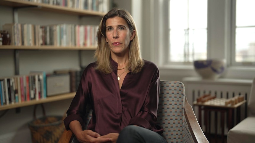 A woman sitting indoors with her hands on her lap. Bookshelves can be seen behind her.