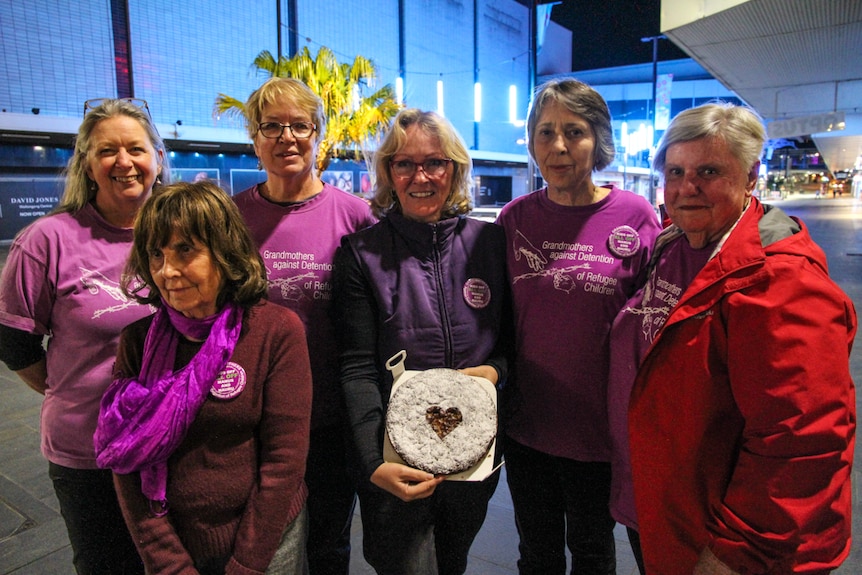 Six women, one of them holding a chocolate cake, standing in the Wollongong Mall at night.