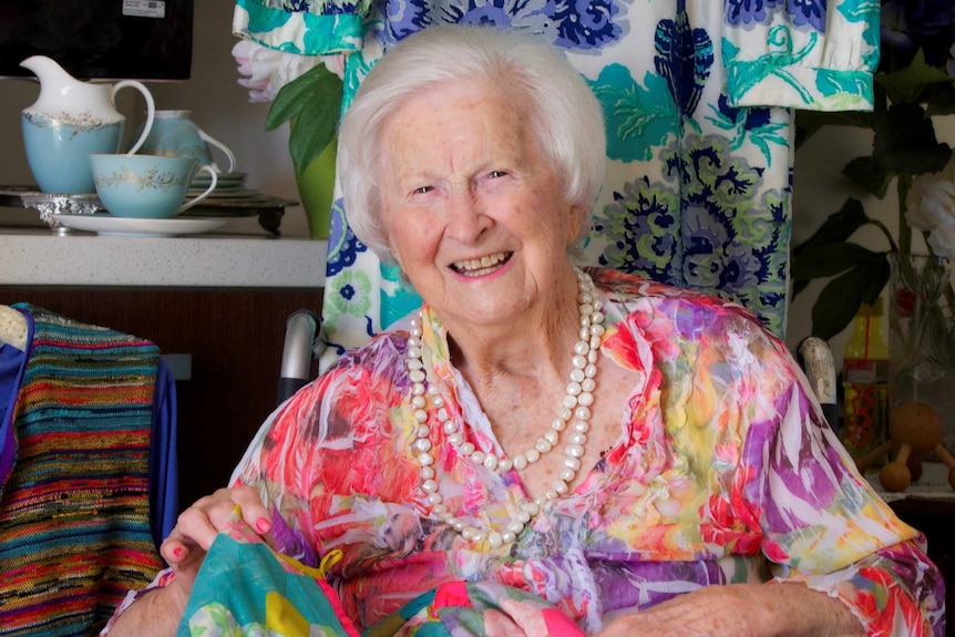 Elderly woman with white hair smiles as she sits in front of a colourful dress, with a colourful jacket over her legs.